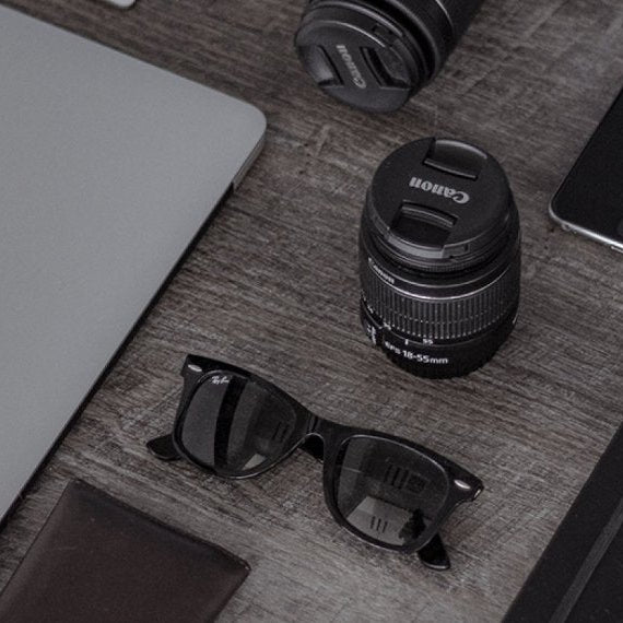 Laptop, camera lens, sunglasses, phone, and wallet on a wooden surface