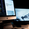 Two computer monitors on a desk with one displaying code and the other showing a mountain landscape.